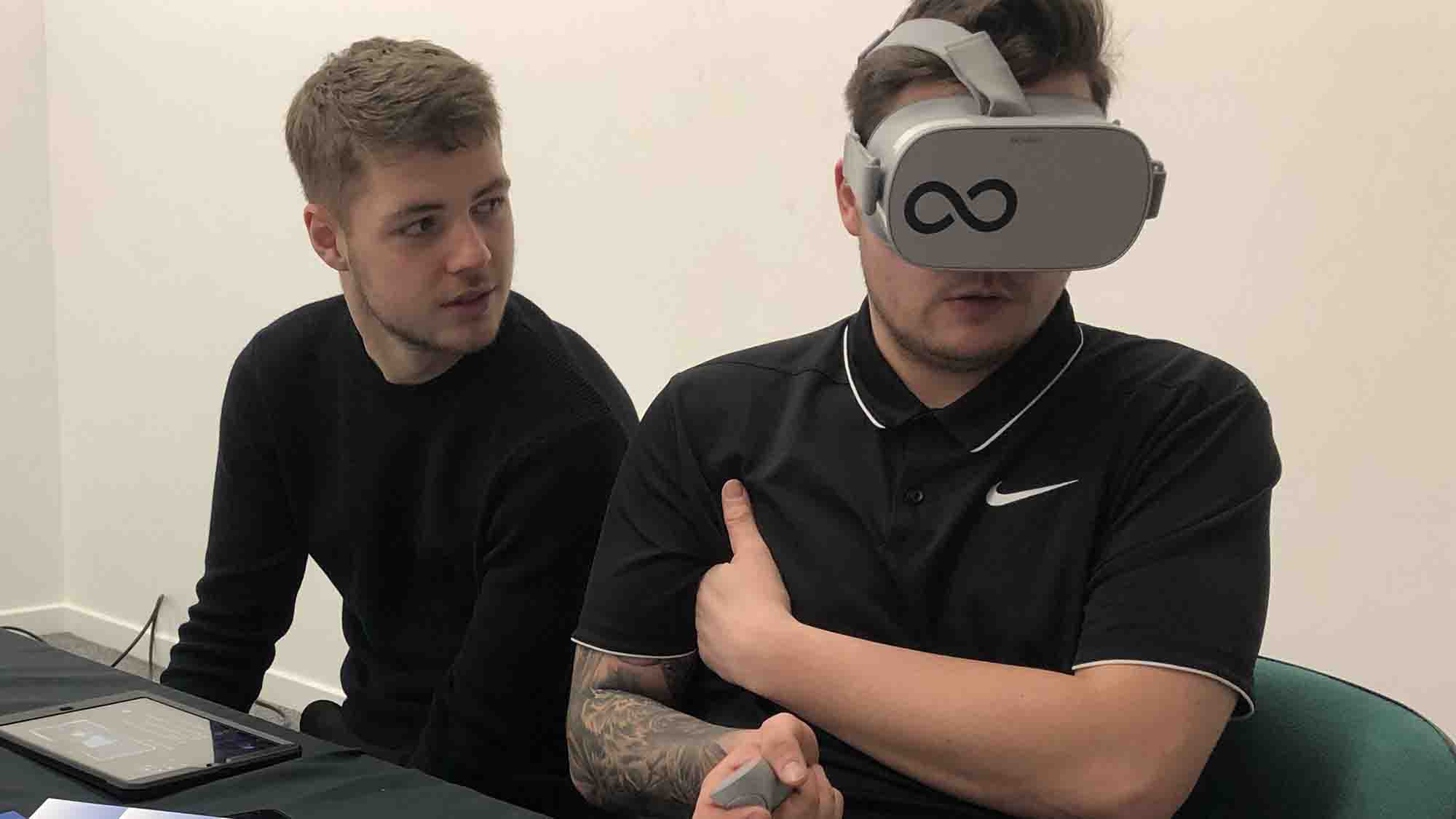 Seated man wearing a VR headset and controller is observed by a colleague during testing.