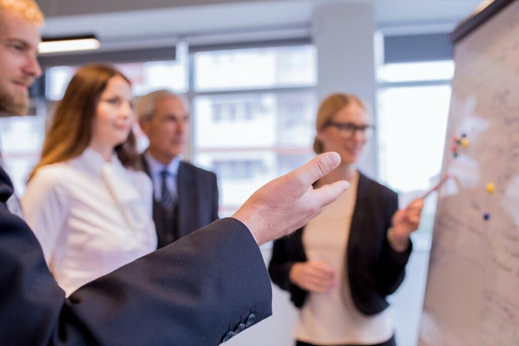 Business team in an office listening to a presenter pointing at a flip chart.