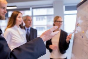 Business team in an office listening to a presenter pointing at a flip chart.